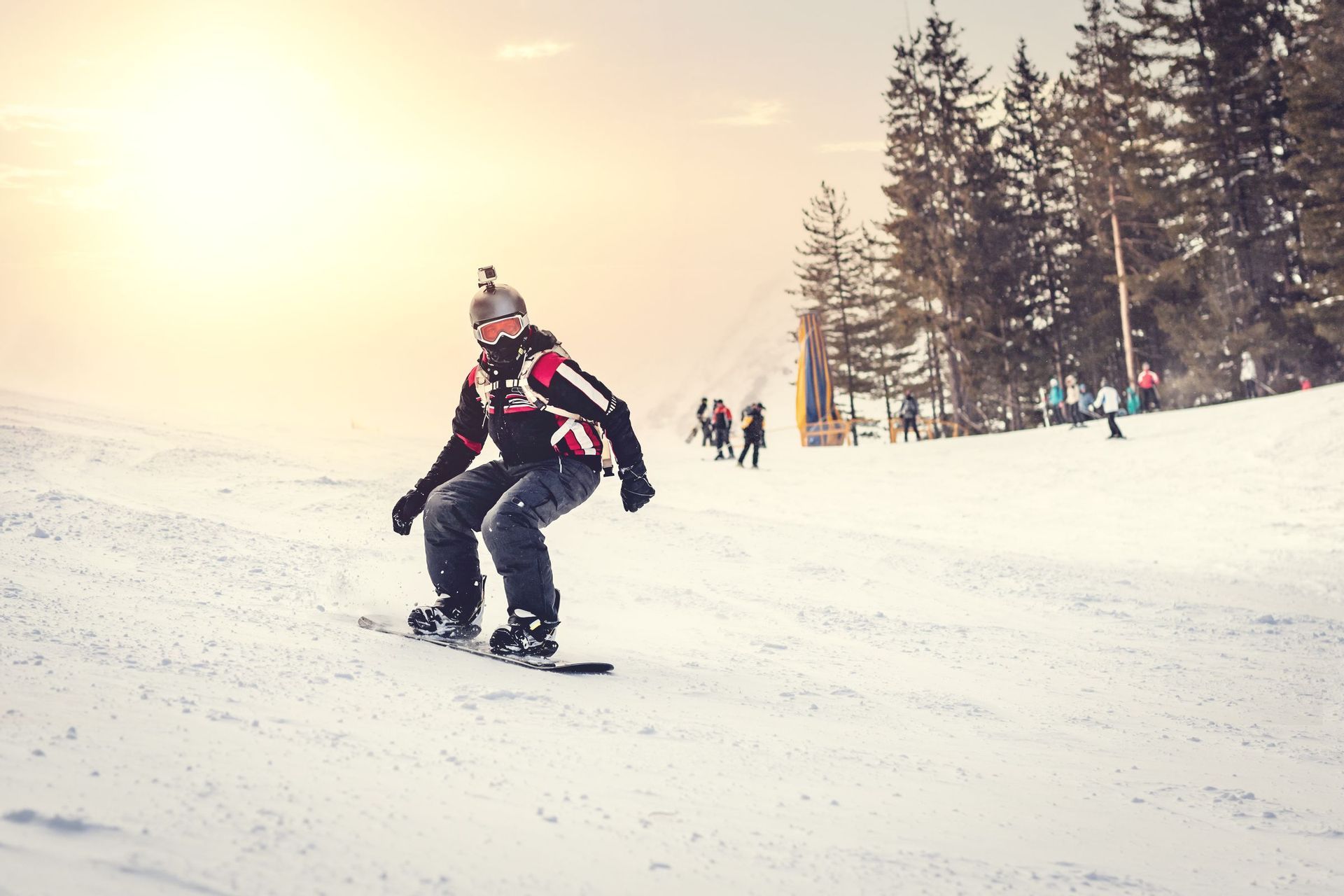 Eine Person mit Helm und Kamera fährt einen verschneiten Hang hinunter, mit Kiefern und anderen Personen im Hintergrund bei Sonnenuntergang.