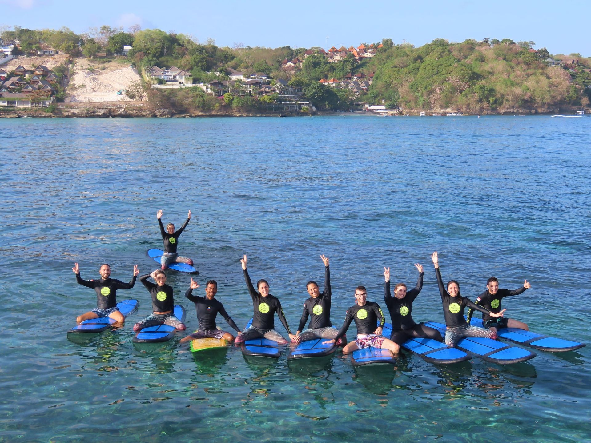 Eine WeRoad Gruppenreise in Neoprenanzügen posiert auf blauen Surfbrettern im klaren Meer, mit einer grünen Küstenlinie im Hintergrund.