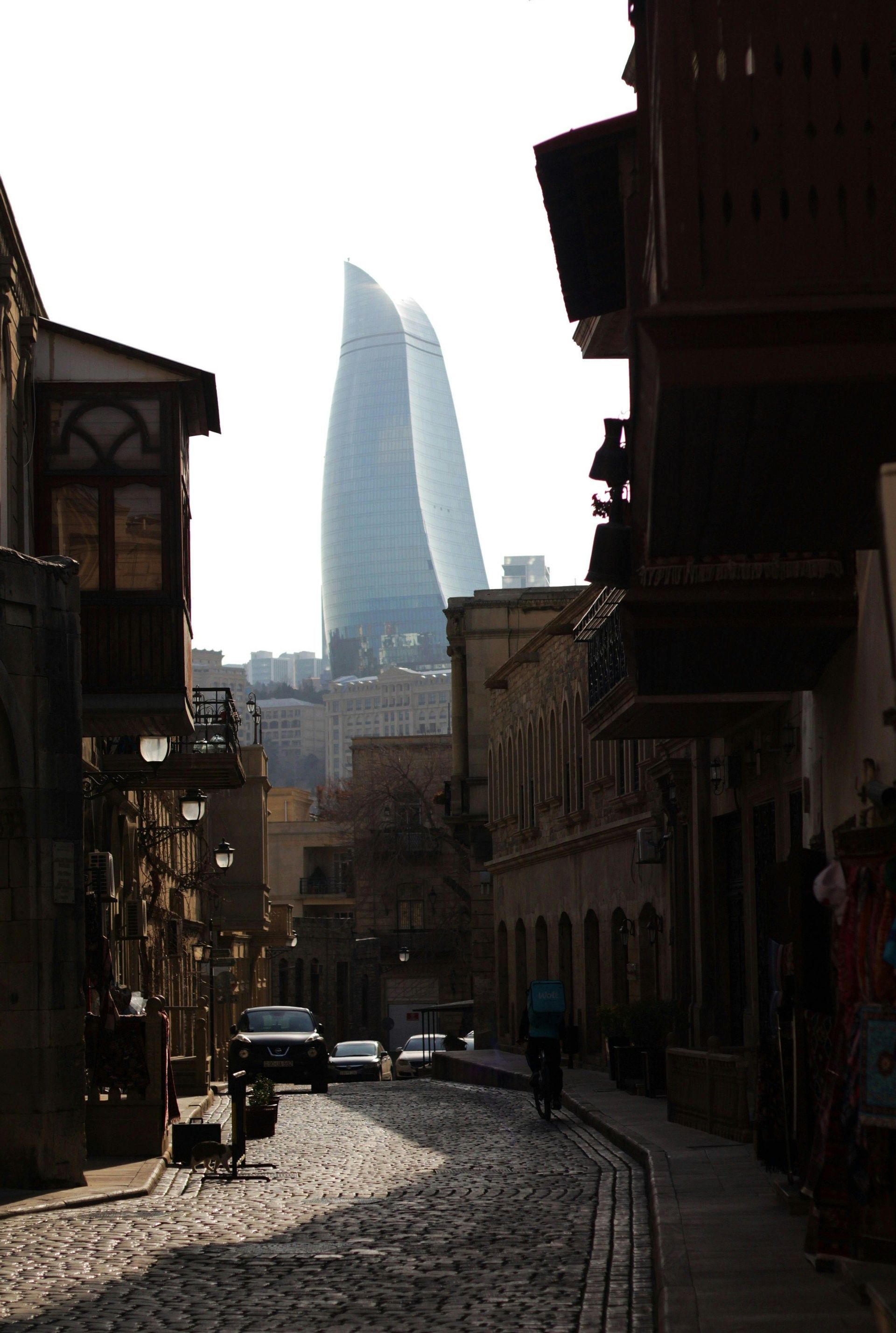 Un moderno grattacielo di vetro si erge su una stretta strada acciottolata fiancheggiata da edifici tradizionali in ombra.