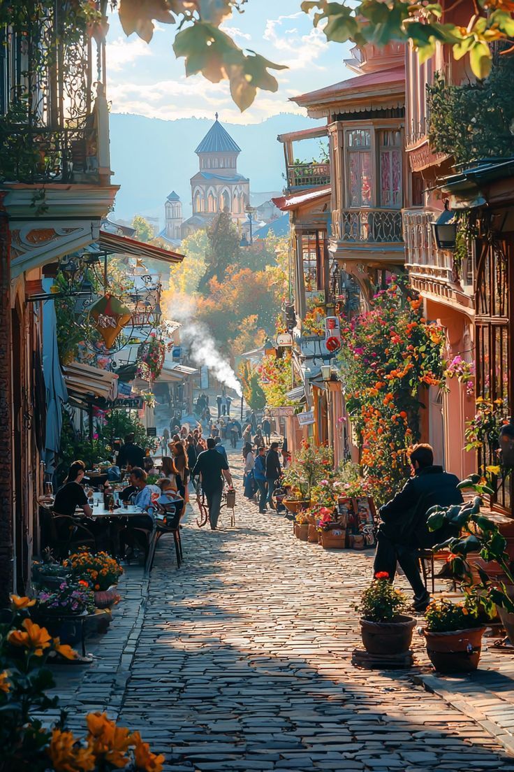 Le persone passeggiano e si siedono nei caffè all'aperto su una strada acciottolata illuminata dal sole, fiancheggiata da fiori ed edifici storici.