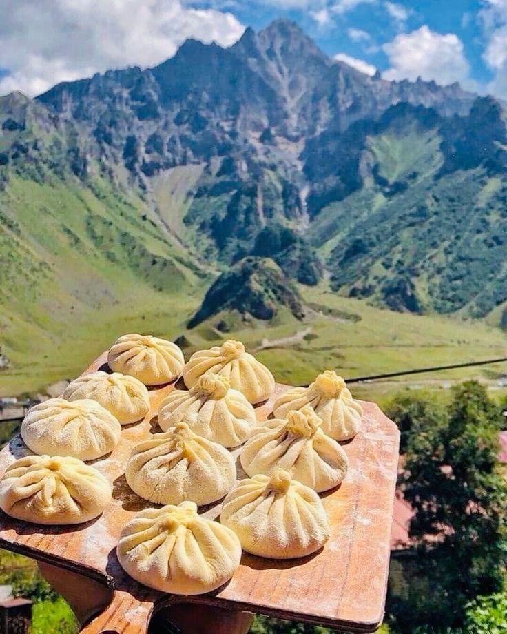 Ravioli pieghettati crudi disposti su un tagliere di legno, sullo sfondo di montagne verdi.