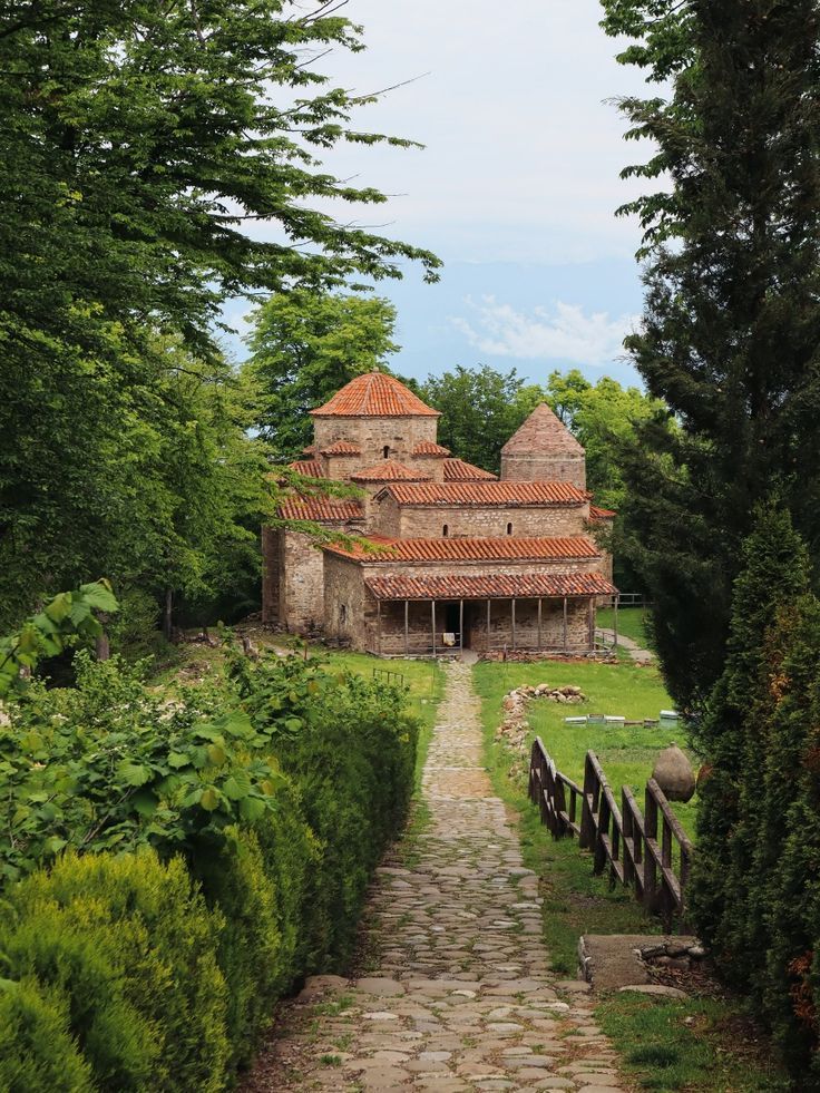 Un rustico monastero in pietra con tetti di tegole rosse sorge alla fine di un sentiero acciottolato, immerso in una lussureggiante vegetazione verde.