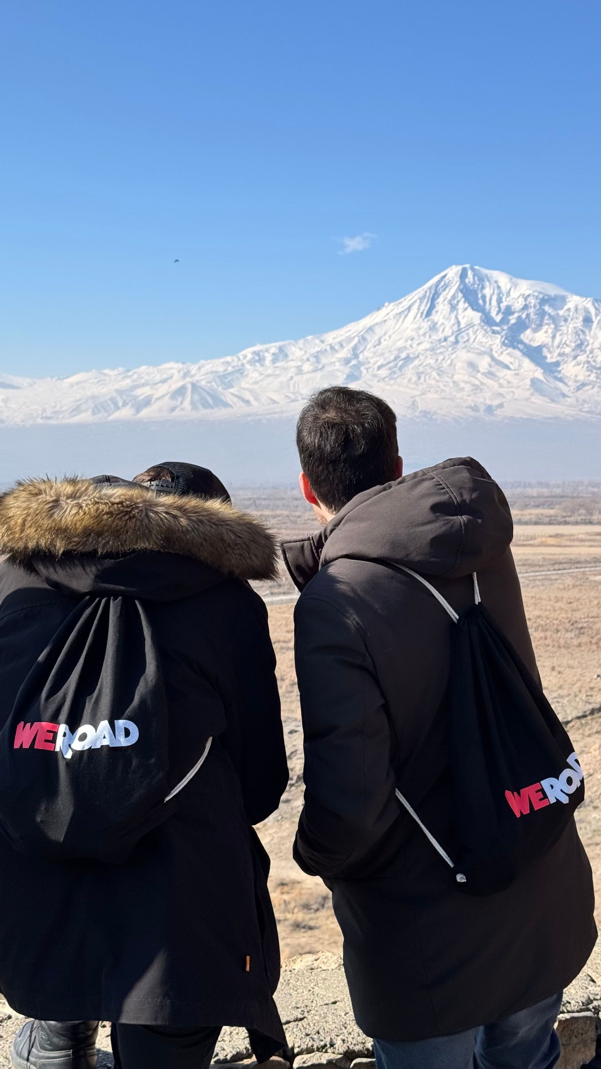 Due persone di un viaggio di gruppo WeRoad, viste di spalle, che guardano una grande montagna innevata sotto un cielo azzurro e limpido.