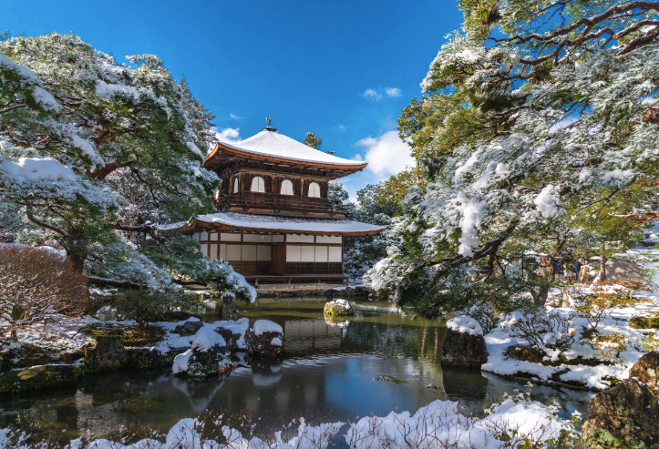 Ein traditioneller japanischer Tempel steht an einem Teich, umgeben von schneebedeckten Bäumen unter klarem blauem Himmel.