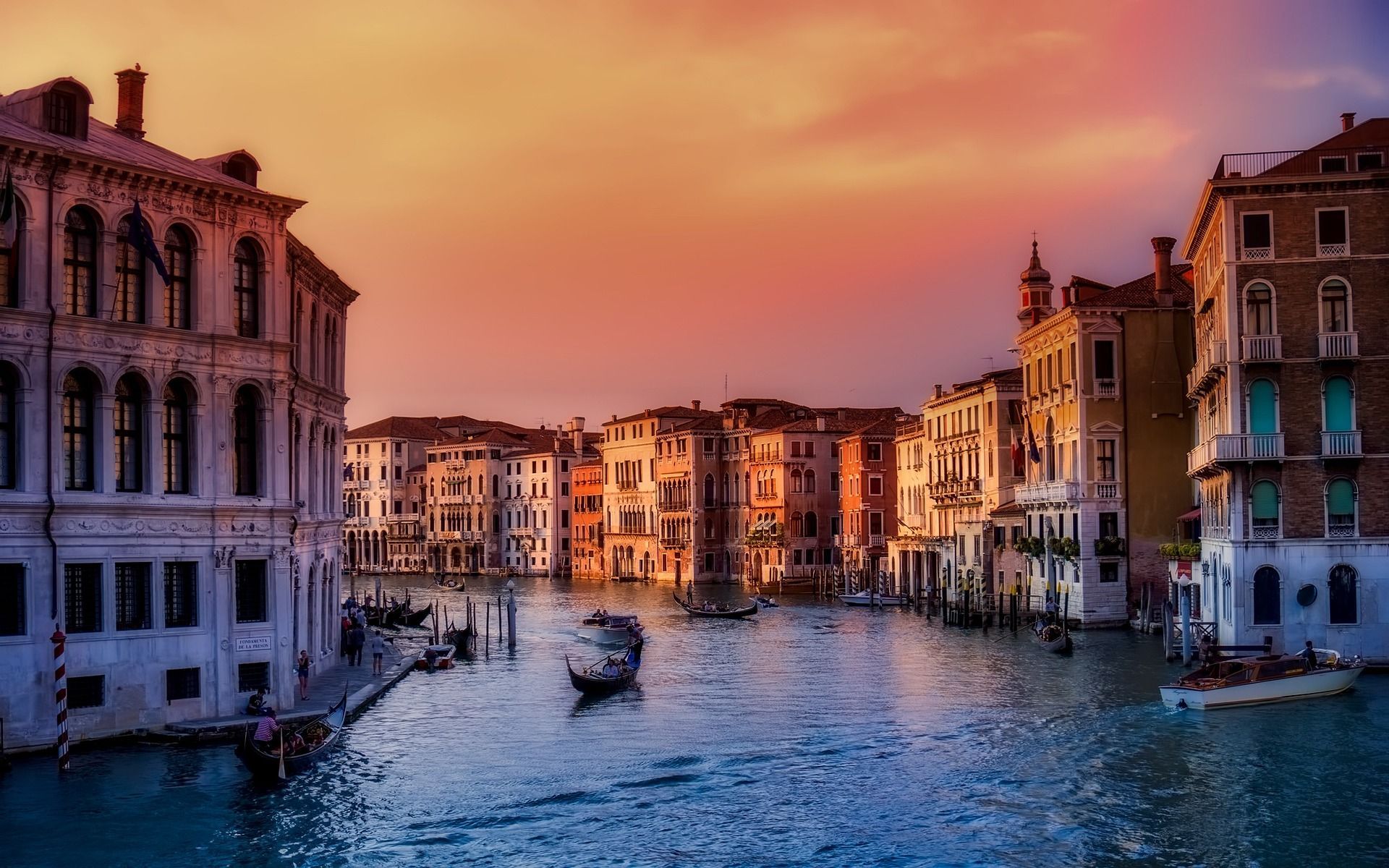 Góndolas y barcos navegan por un ancho canal flanqueado por edificios históricos bajo un cielo de atardecer colorido.