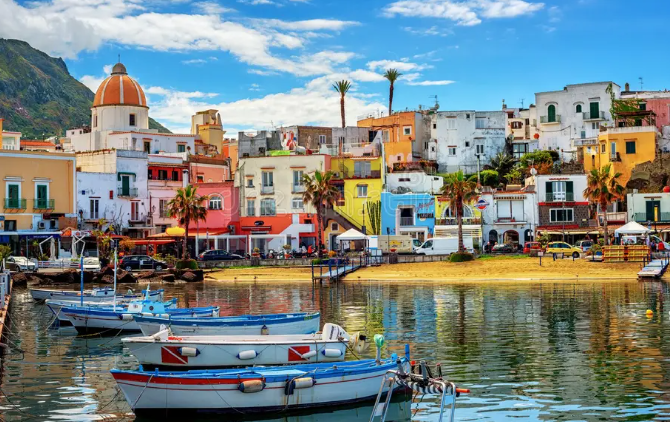 Piccole imbarcazioni sono ormeggiate in un porto di fronte a una colorata città costiera con una spiaggia sabbiosa e una chiesa a cupola.