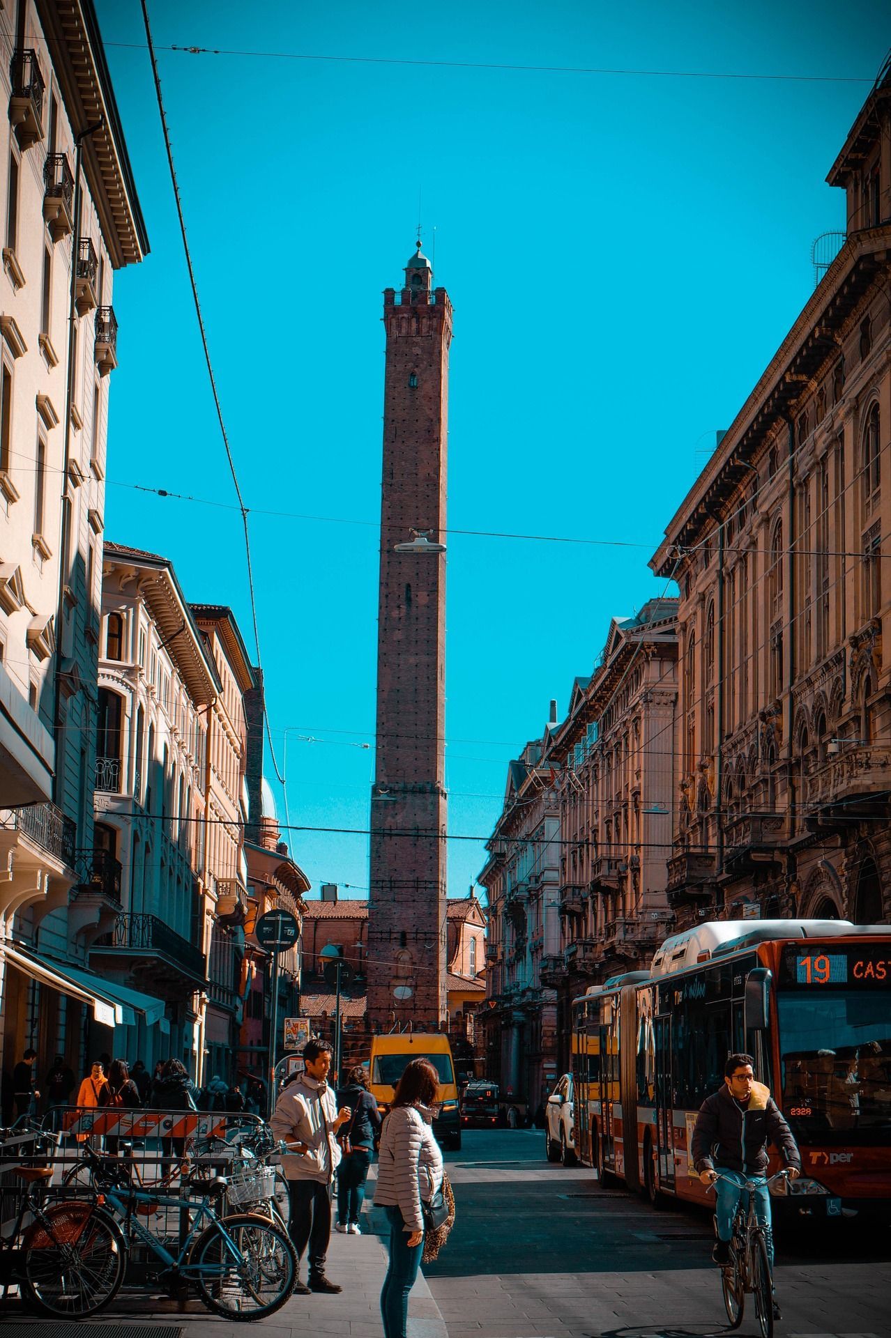Una alta torre de ladrillo se alza al final de una estrecha calle de la ciudad con gente, bicicletas y un autobús bajo un cielo azul claro.