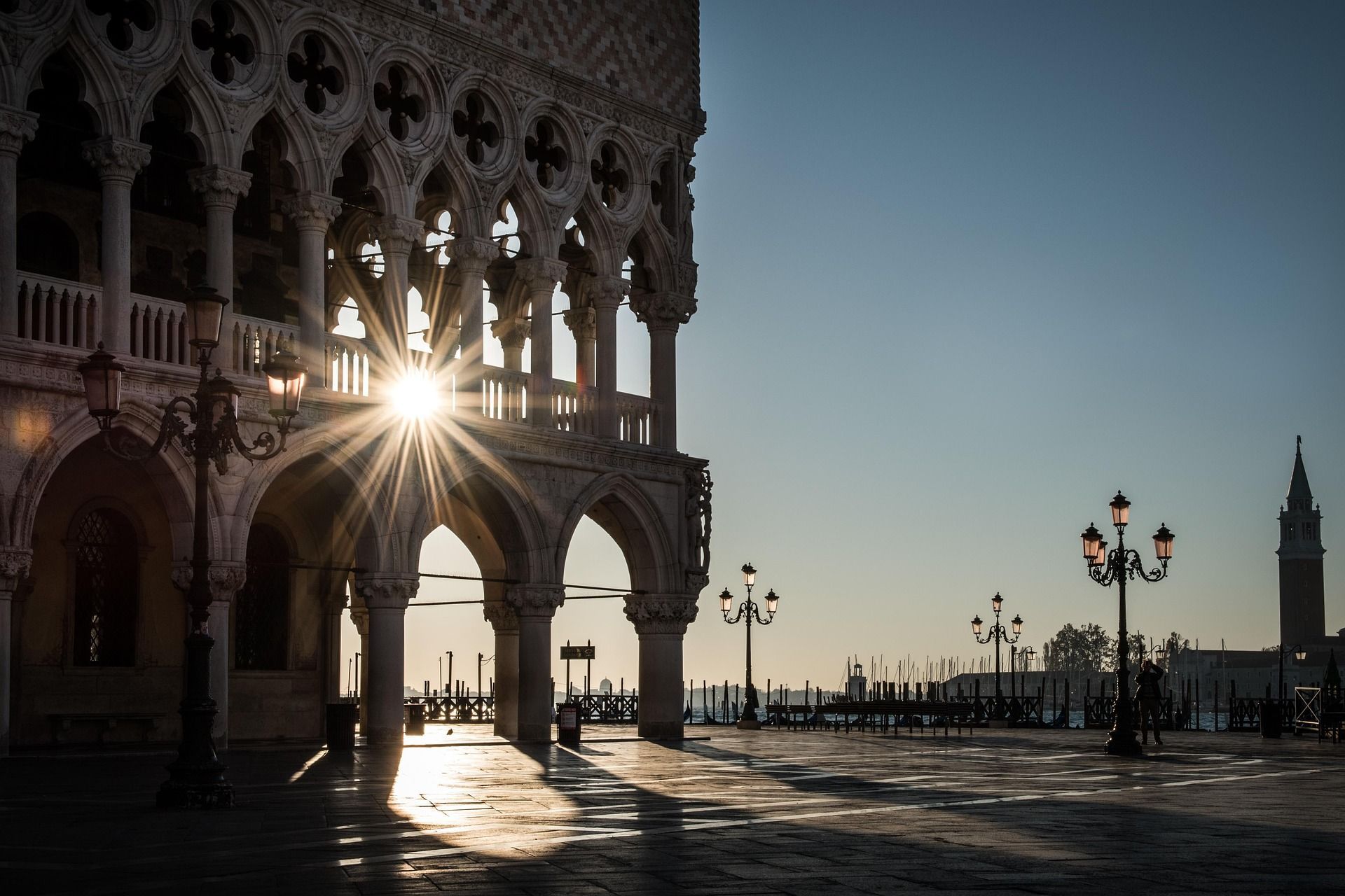 Un brillante rayo de sol atraviesa los arcos de un gran edificio, proyectando largas sombras sobre una plaza pavimentada vacía.