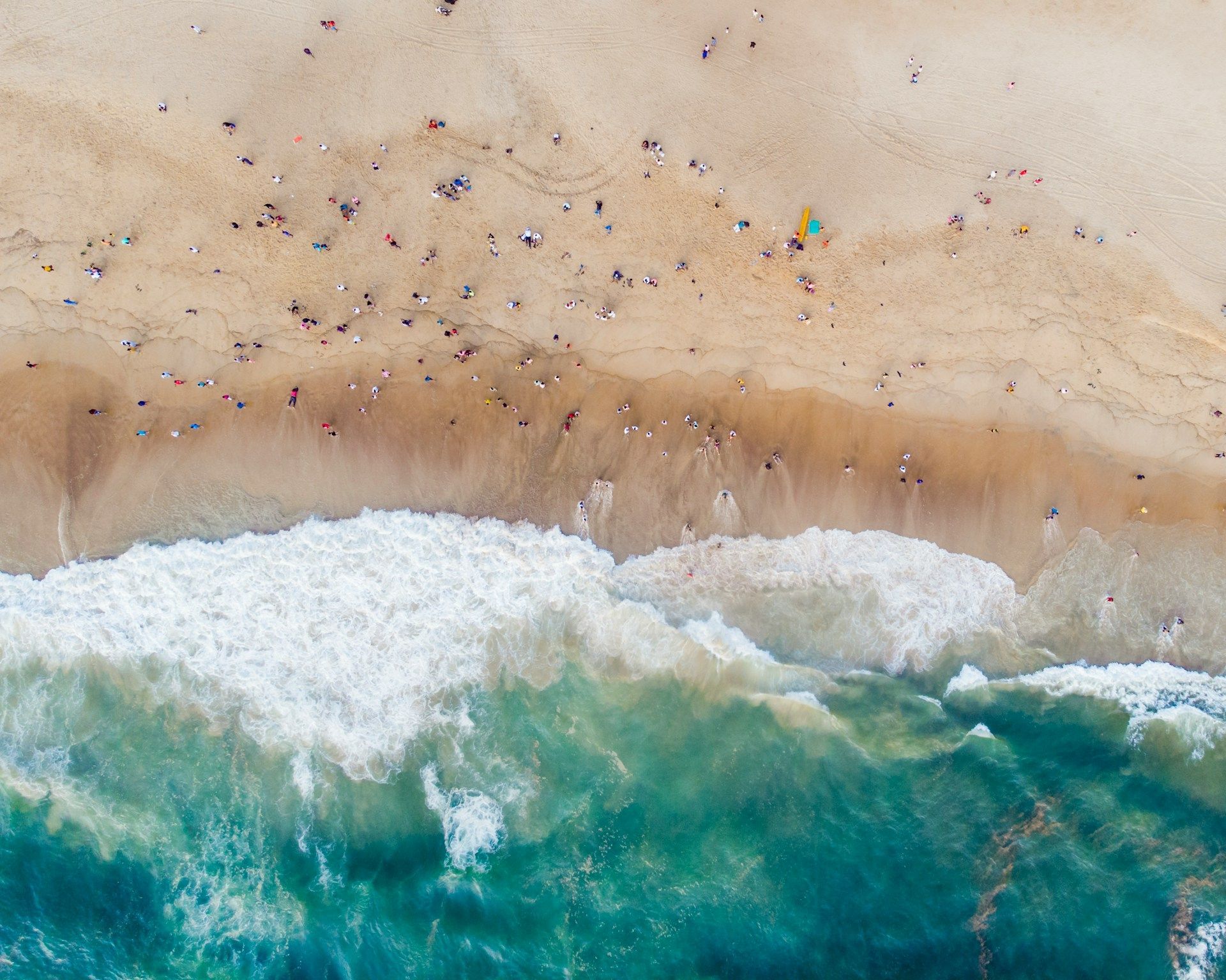 Una vista aerea dall'alto di una spiaggia sabbiosa affollata con persone, mentre onde turchesi dell'oceano con schiuma bianca si infrangono a riva.