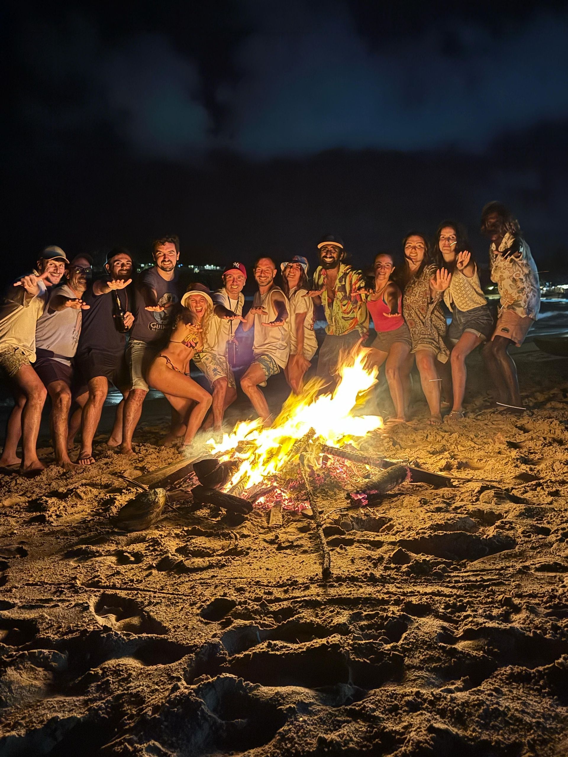 Un viaggio di gruppo WeRoad in posa attorno a un grande falò su una spiaggia sabbiosa di notte.
