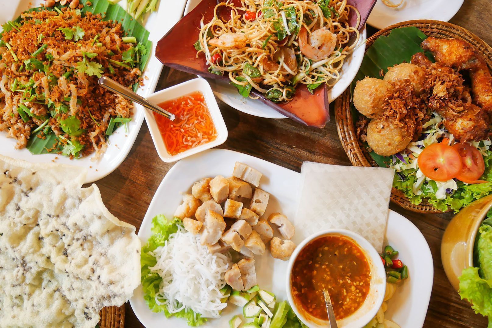 An overhead view of a spread of Asian dishes, including noodle salad with shrimp, fried items, and fresh vegetables on a wooden table.