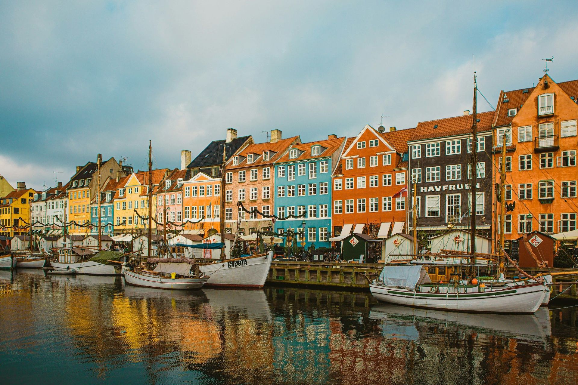 A row of colorful buildings and docked sailboats reflected in a canal under a cloudy sky.