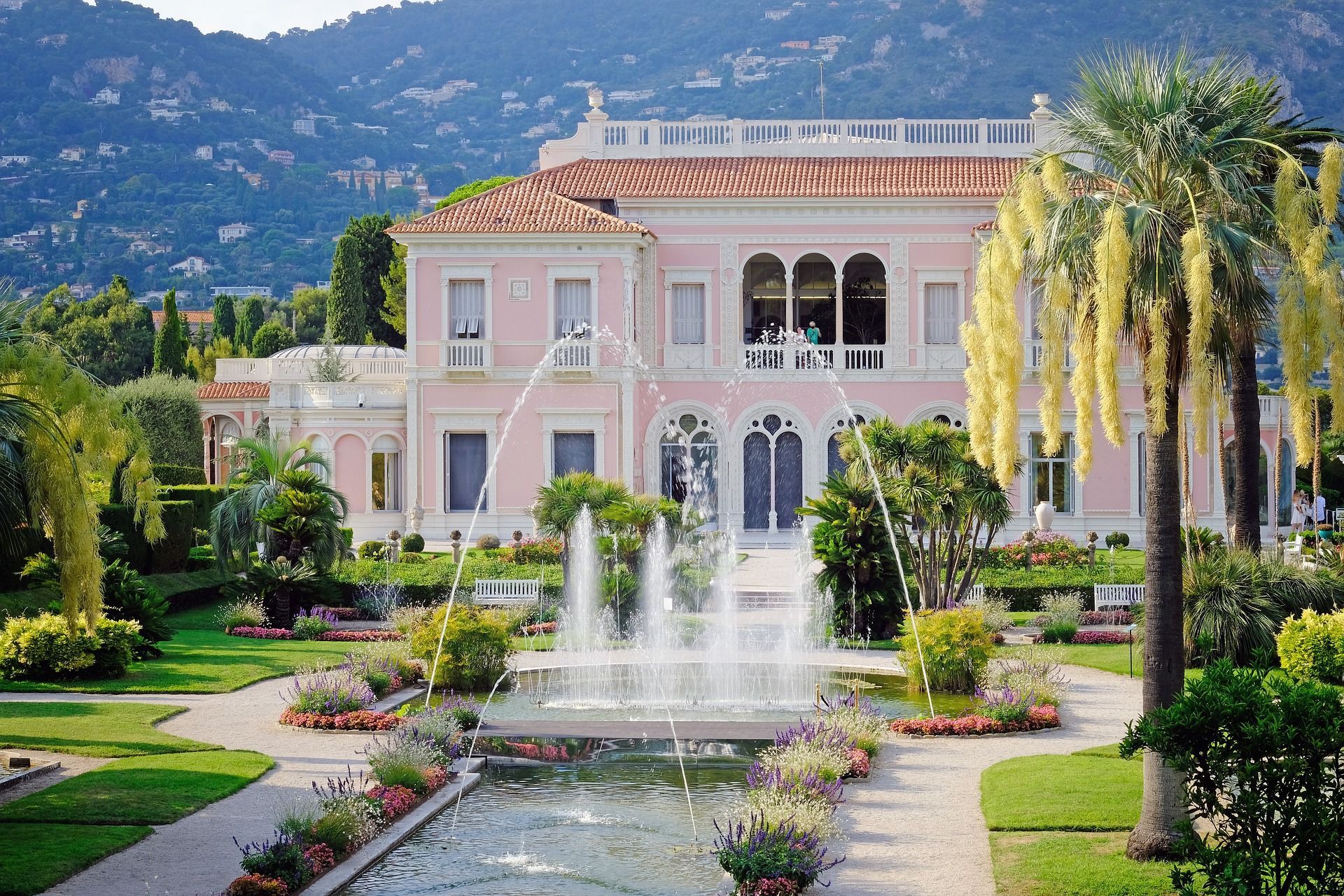 Una gran villa rosa con fuentes que rocían agua en un jardín impecable, con palmeras y una montaña al fondo.