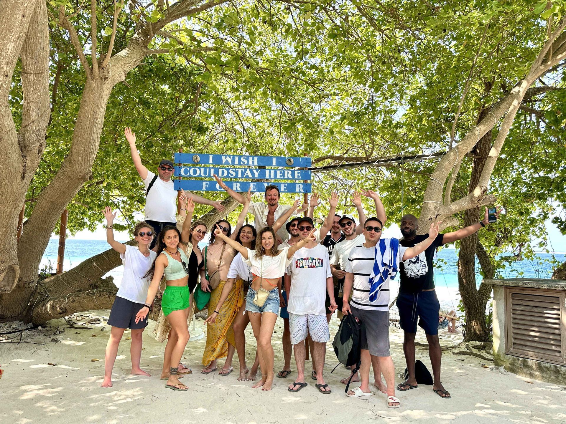 Un groupe WeRoad posant pour une photo sur une plage de sable sous un grand arbre feuillu avec un panneau bleu.