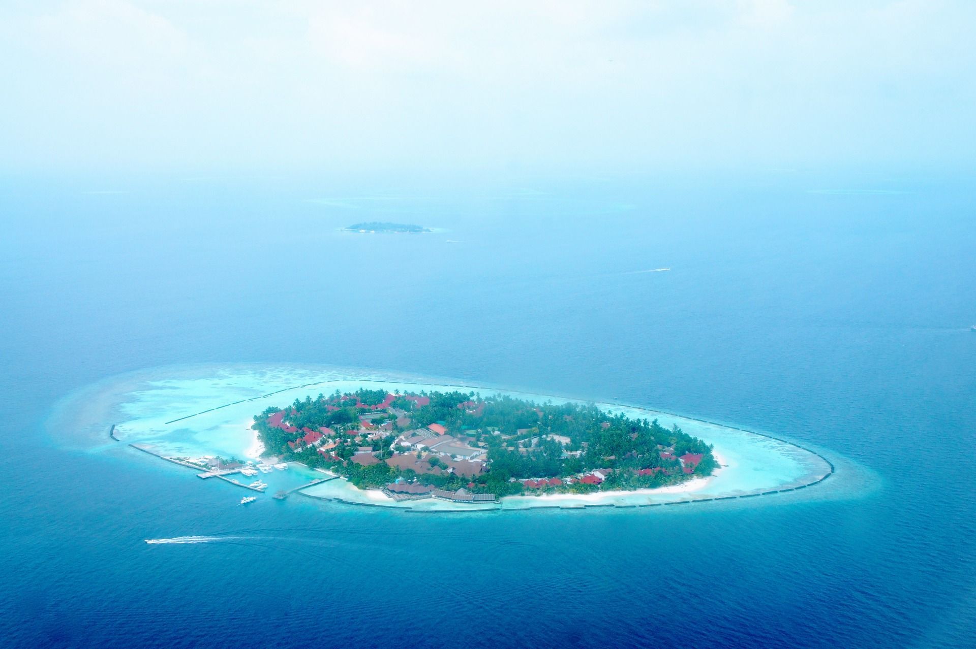 Vue aérienne d'un petit complexe hôtelier tropical sur une île, avec des palmiers, entouré d'un lagon turquoise et d'un océan bleu profond.
