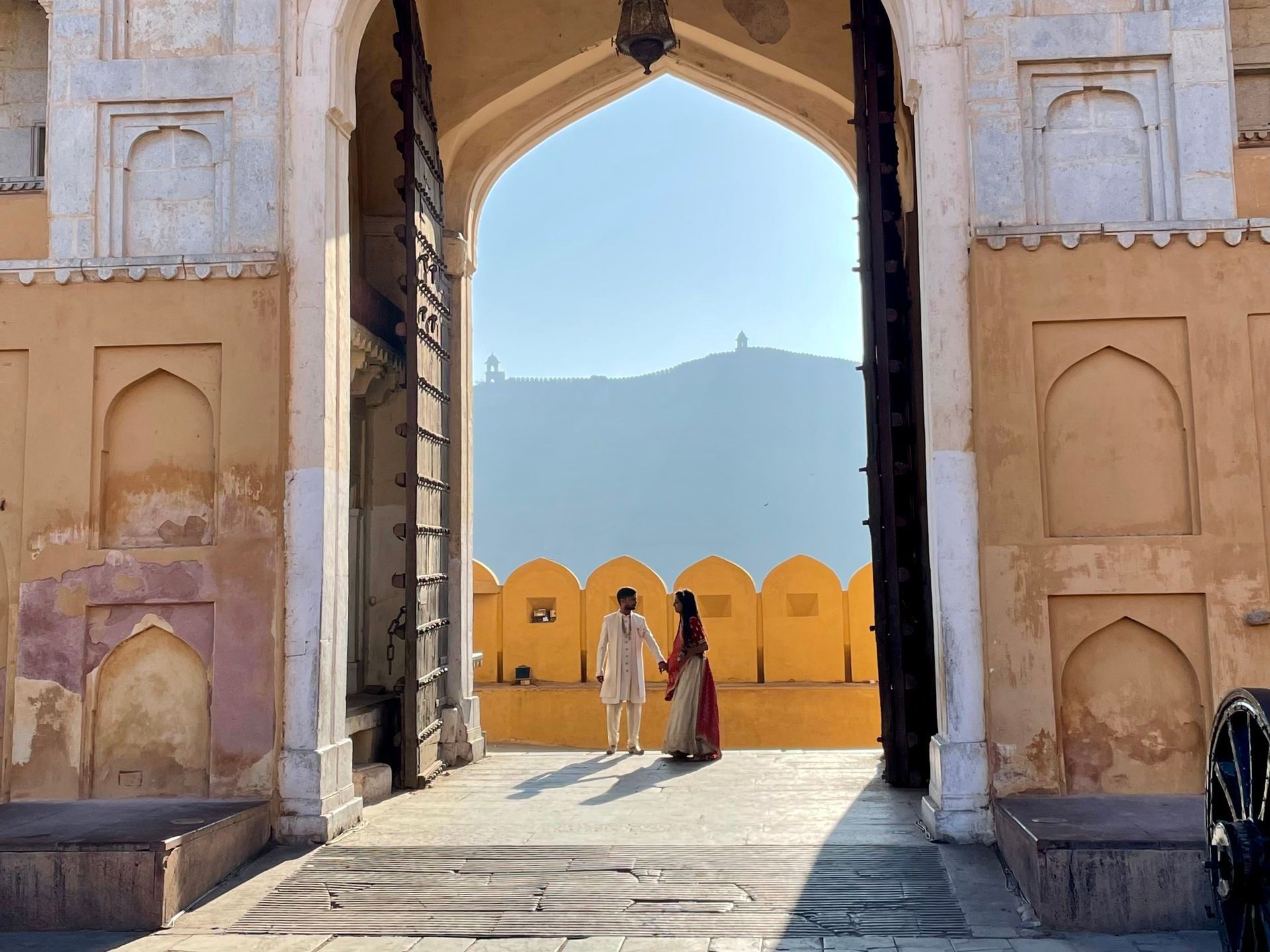 Un couple en tenue indienne traditionnelle se tient sous une grande arche, avec une colline lointaine visible à travers l'ouverture.