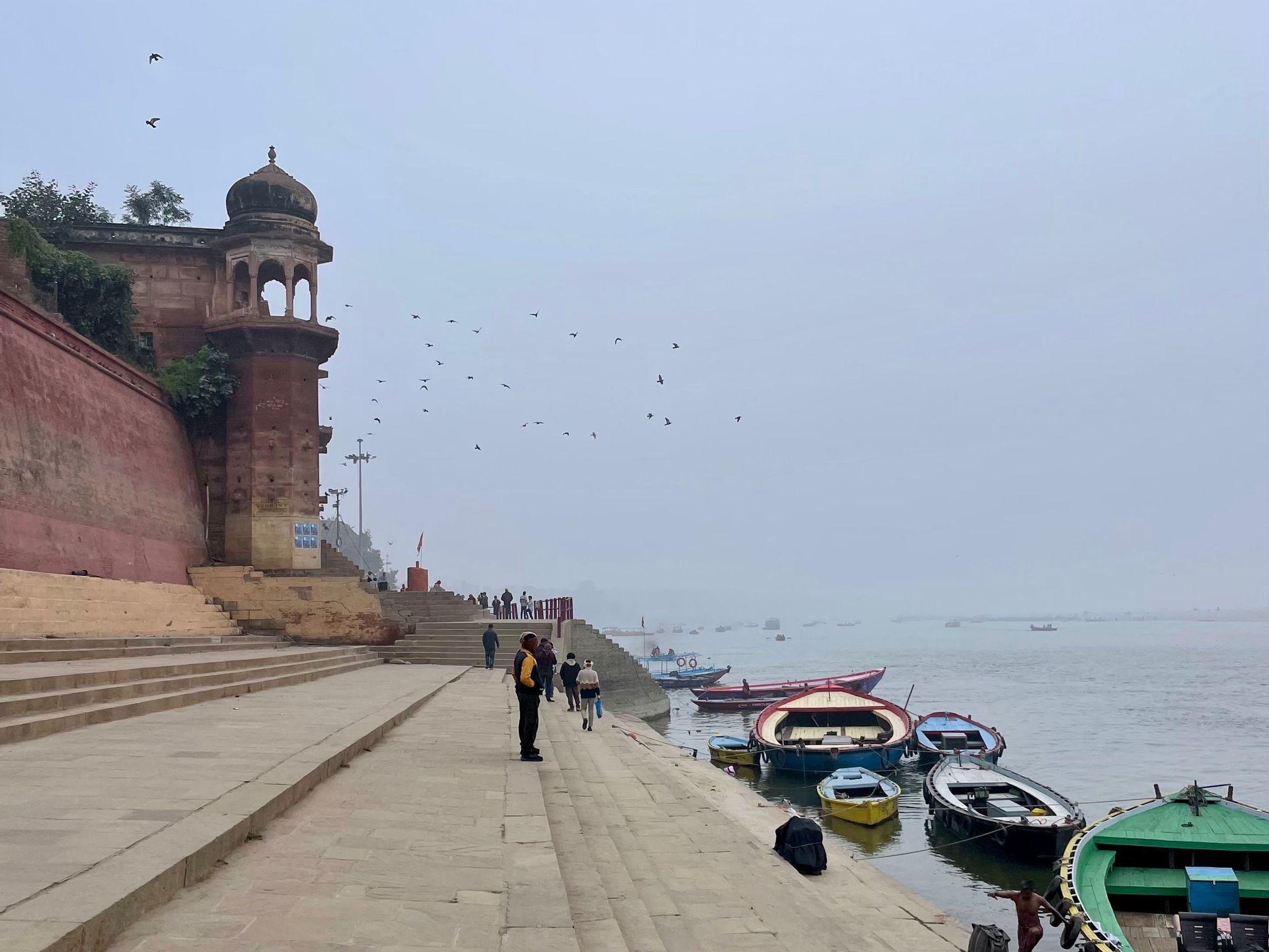 Steinerne Ghats führen hinunter zu einem Fluss, wo bunte Boote vertäut sind und an einem nebligen Tag Menschen vorbeigehen.