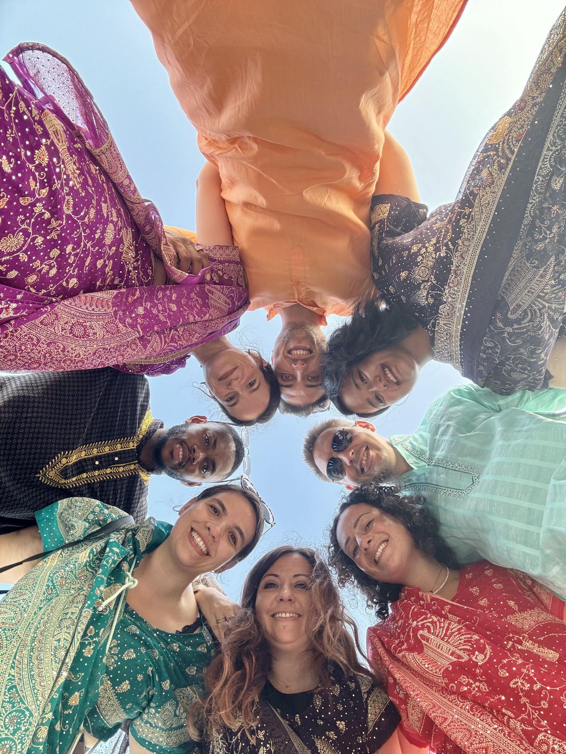Un groupe WeRoad en voyage, portant des vêtements traditionnels colorés, se rassemble en cercle, souriant à l'appareil photo sous un ciel clair.