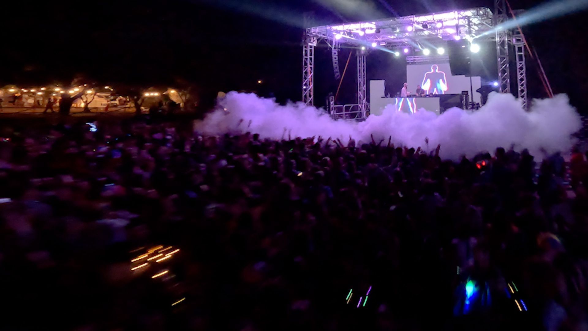 Cover-Mobile.svg Una grande folla balla a un concerto notturno davanti a un palco con un DJ, luci viola ed effetti di fumo.
