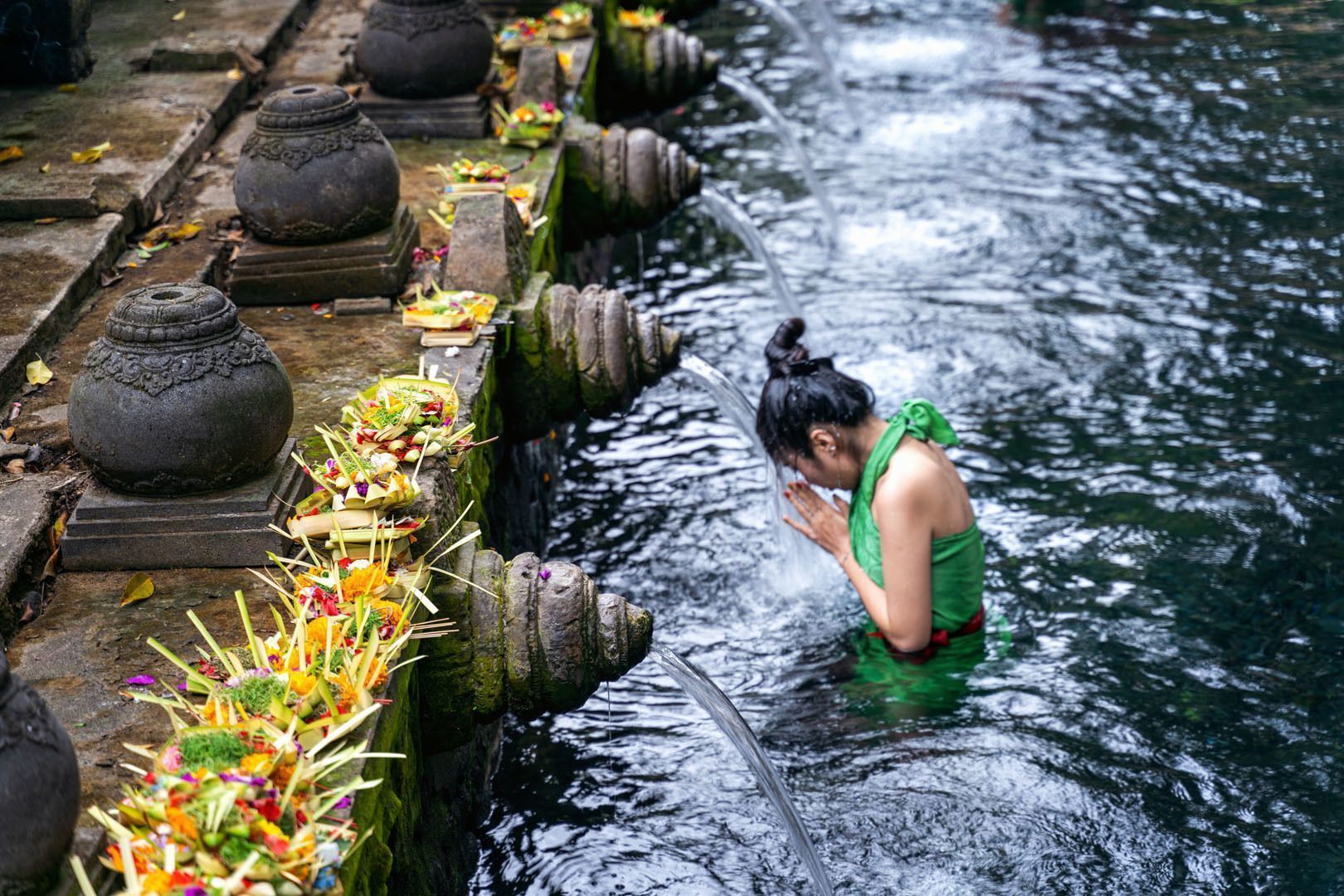 Una mujer con vestimenta verde realiza un ritual de purificación, inclinándose bajo el agua que emana de un caño de piedra en una poza sagrada.
