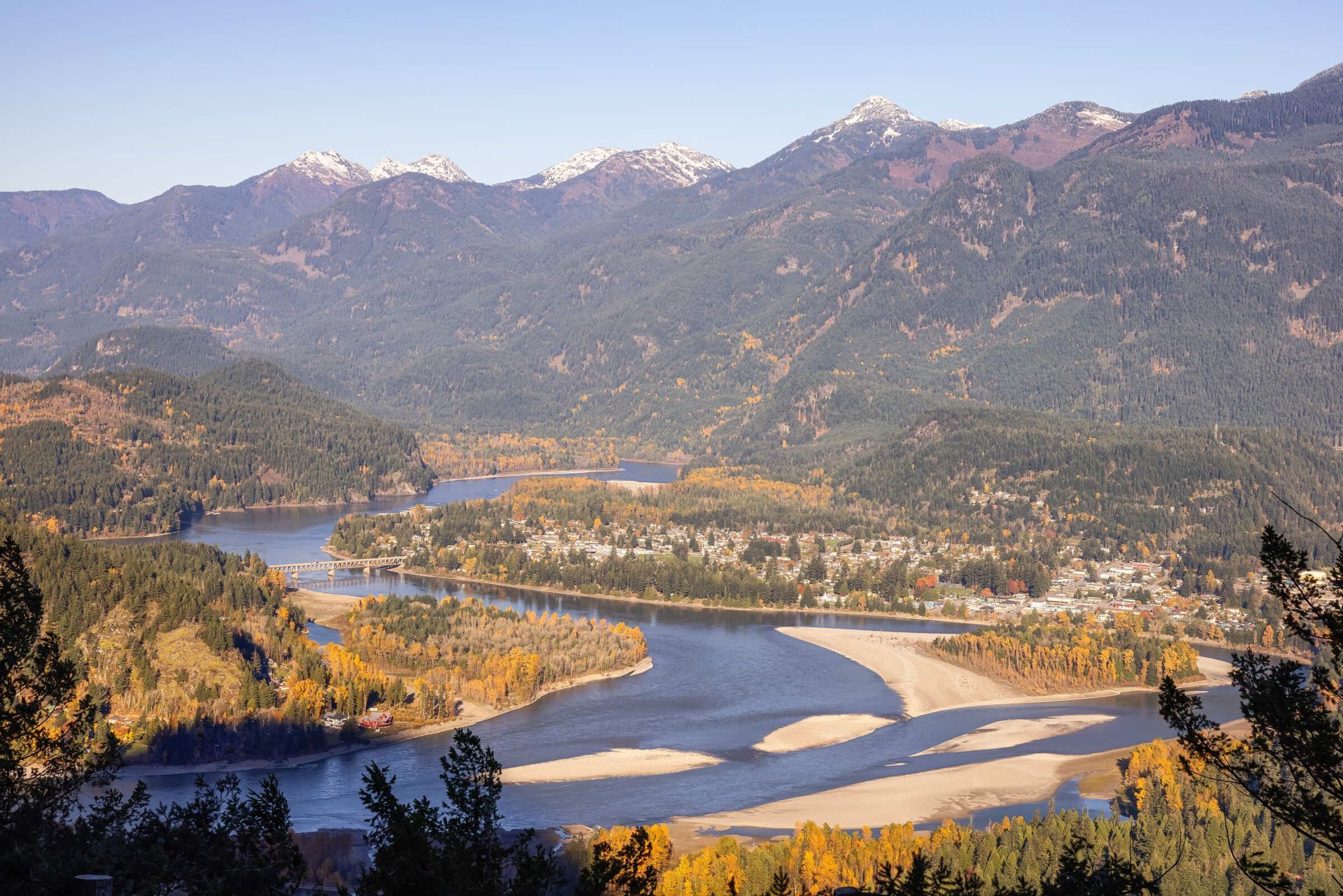 Luftaufnahme eines breiten Flusses mit Sandbänken, der durch ein Herbsttal fließt, mit einer kleinen Stadt und schneebedeckten Bergen.