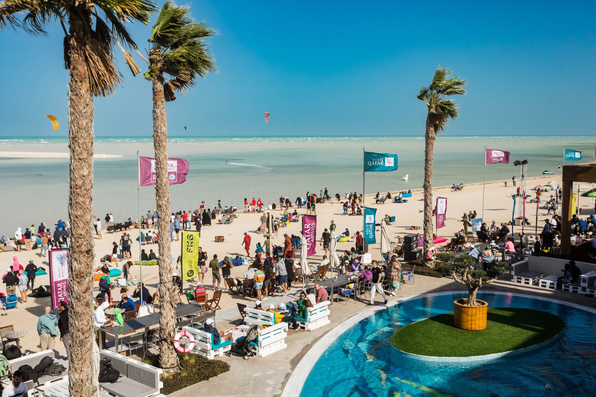 Vista dall'alto di un affollato resort balneare durante un festival soleggiato, con piscina, palme e kitesurfer sul mare.