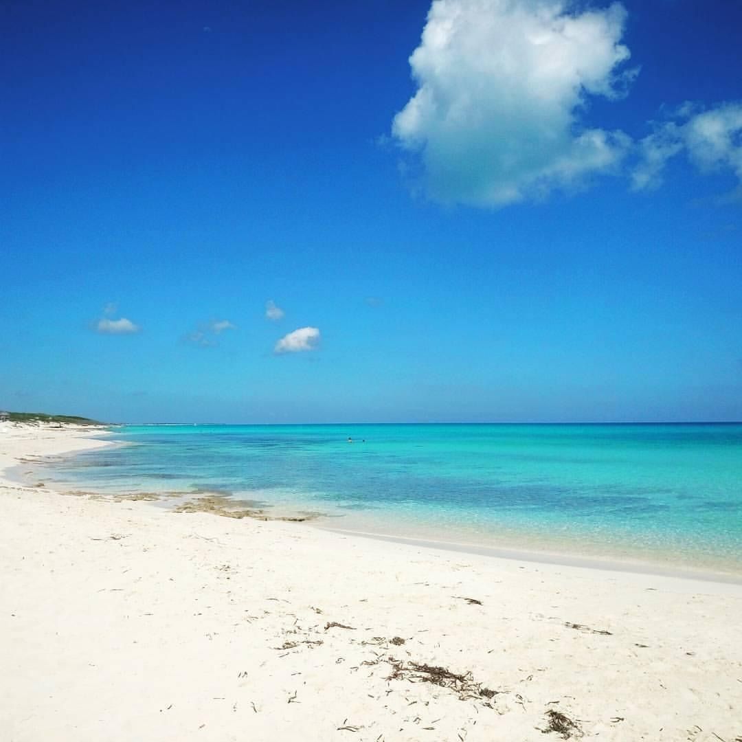 Una spiaggia di sabbia bianca si curva lungo acque calme e turchesi, sotto un cielo azzurro brillante con poche nuvole bianche.