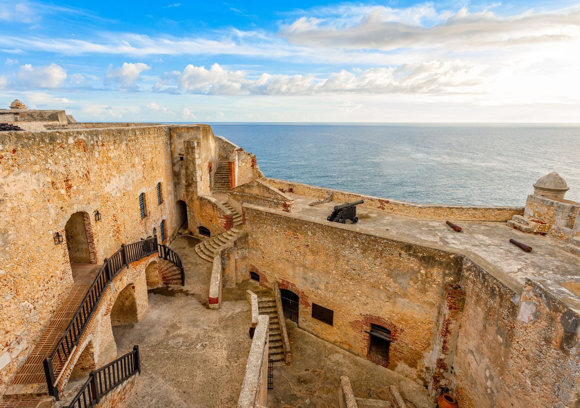 Una fortezza costiera in pietra, a più livelli, con cannoni sulle merlature che domina l'oceano sotto un cielo parzialmente nuvoloso.
