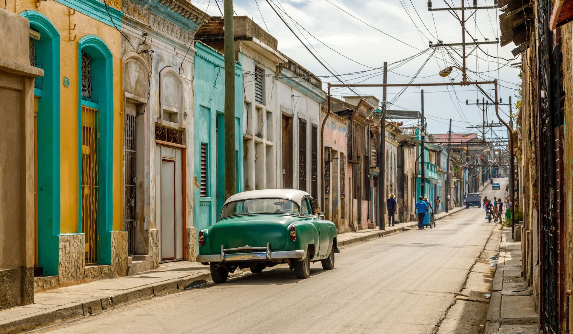 Un'auto d'epoca verde parcheggiata su una strada fiancheggiata da edifici colorati e vissuti e fili elettrici che si incrociano.