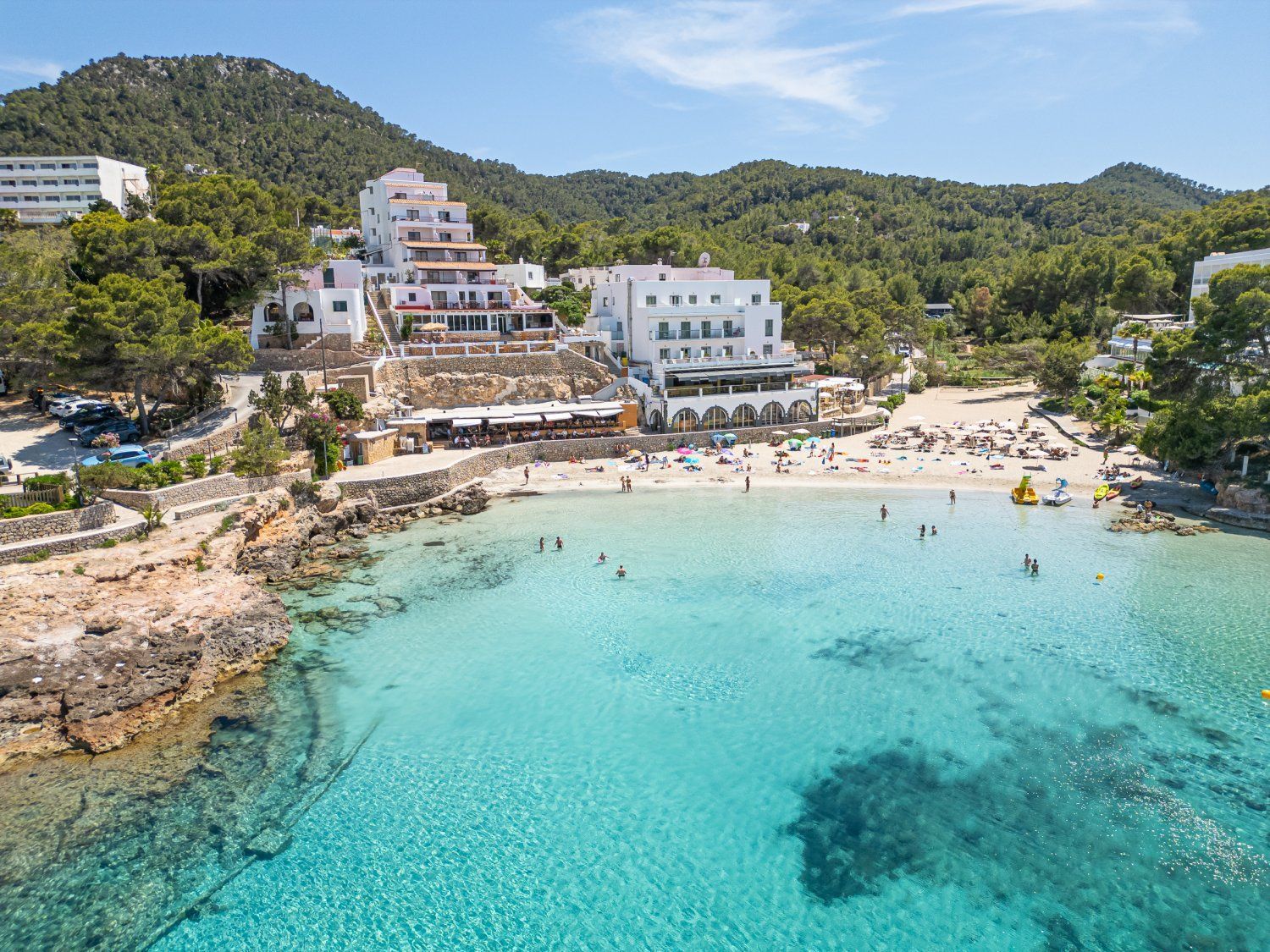 Vista aérea de una cala de playa con gente nadando en agua turquesa y tomando el sol en la arena, respaldada por hoteles blancos en una ladera verde y arbolada.