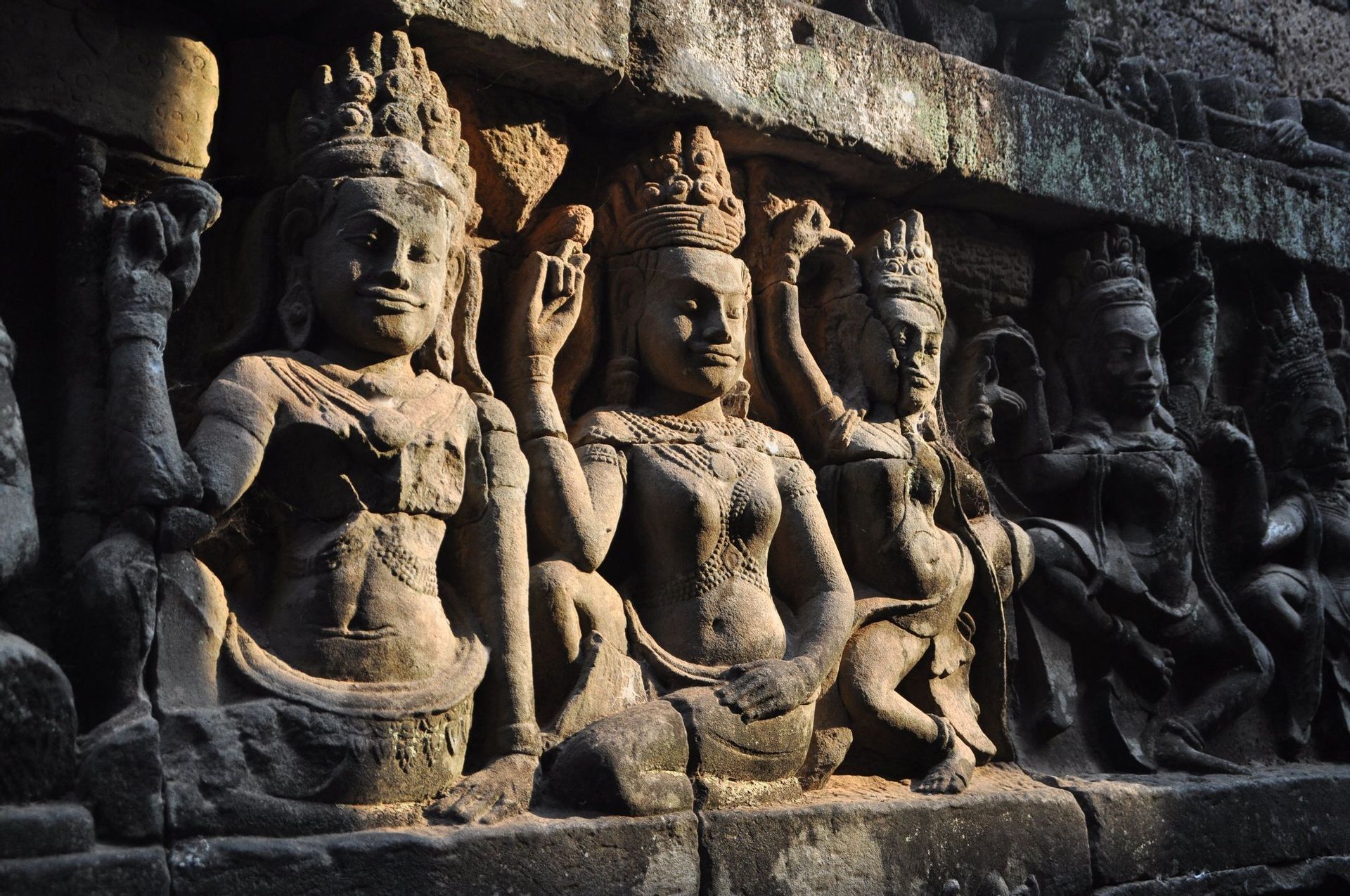 Une rangée de sculptures en pierre anciennes représentant des figures avec des couronnes élaborées et des bijoux sur un mur de temple, éclairée par la lumière du soleil.