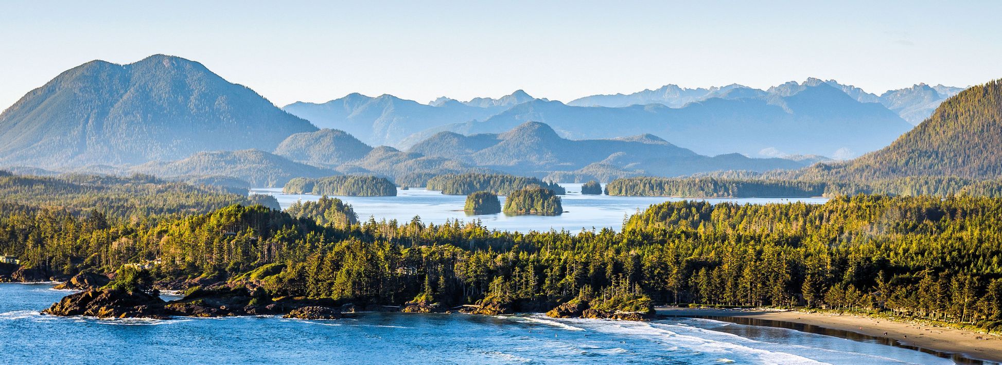 Une vue panoramique d'un littoral avec une plage de sable, un océan bleu et une forêt dense de conifères, avec des îles et des montagnes en arrière-plan.