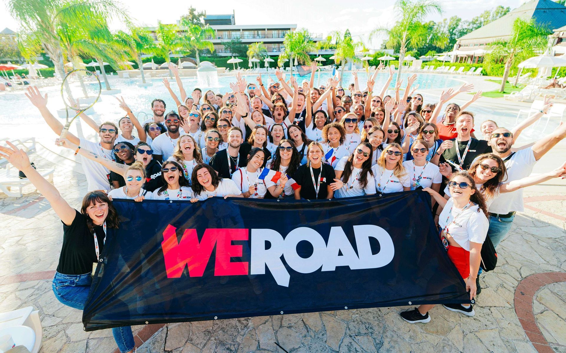 Un grande gruppo WeRoad in viaggio che posa ed esulta per una foto con uno striscione davanti a una piscina.