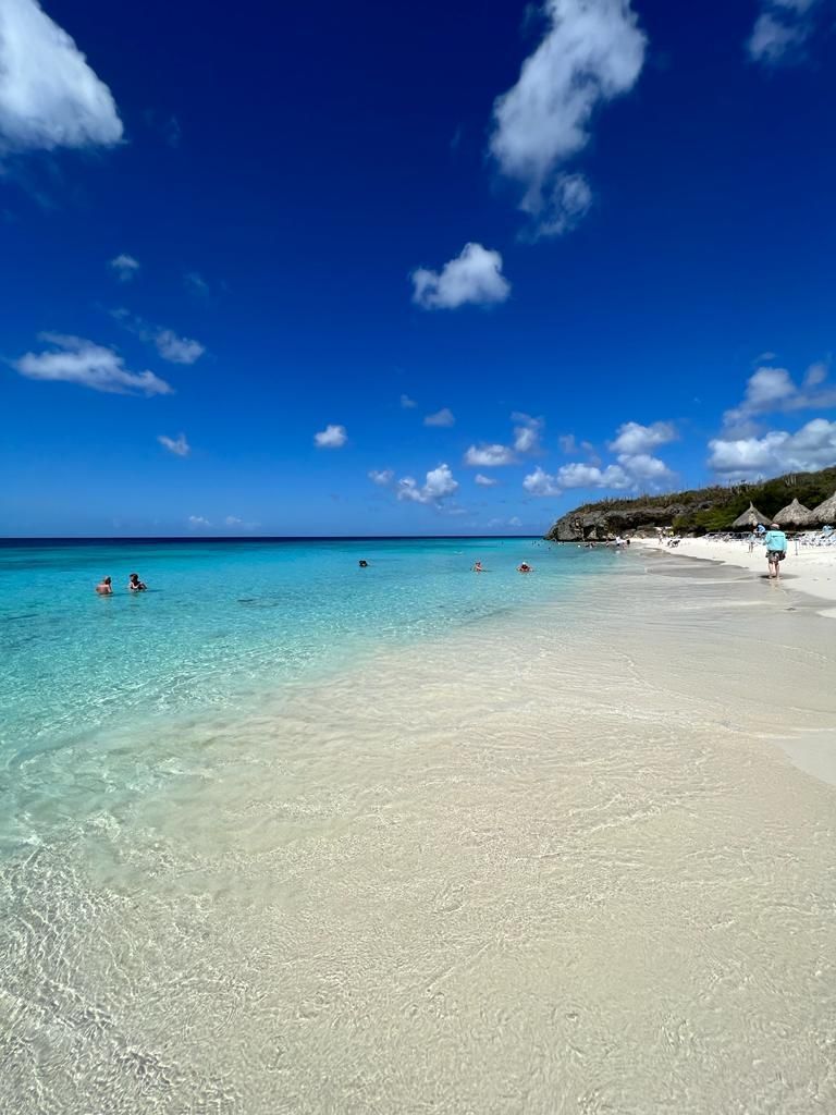 Persone che nuotano in acqua cristallina turchese su una spiaggia di sabbia bianca sotto un cielo blu intenso con nuvole bianche.