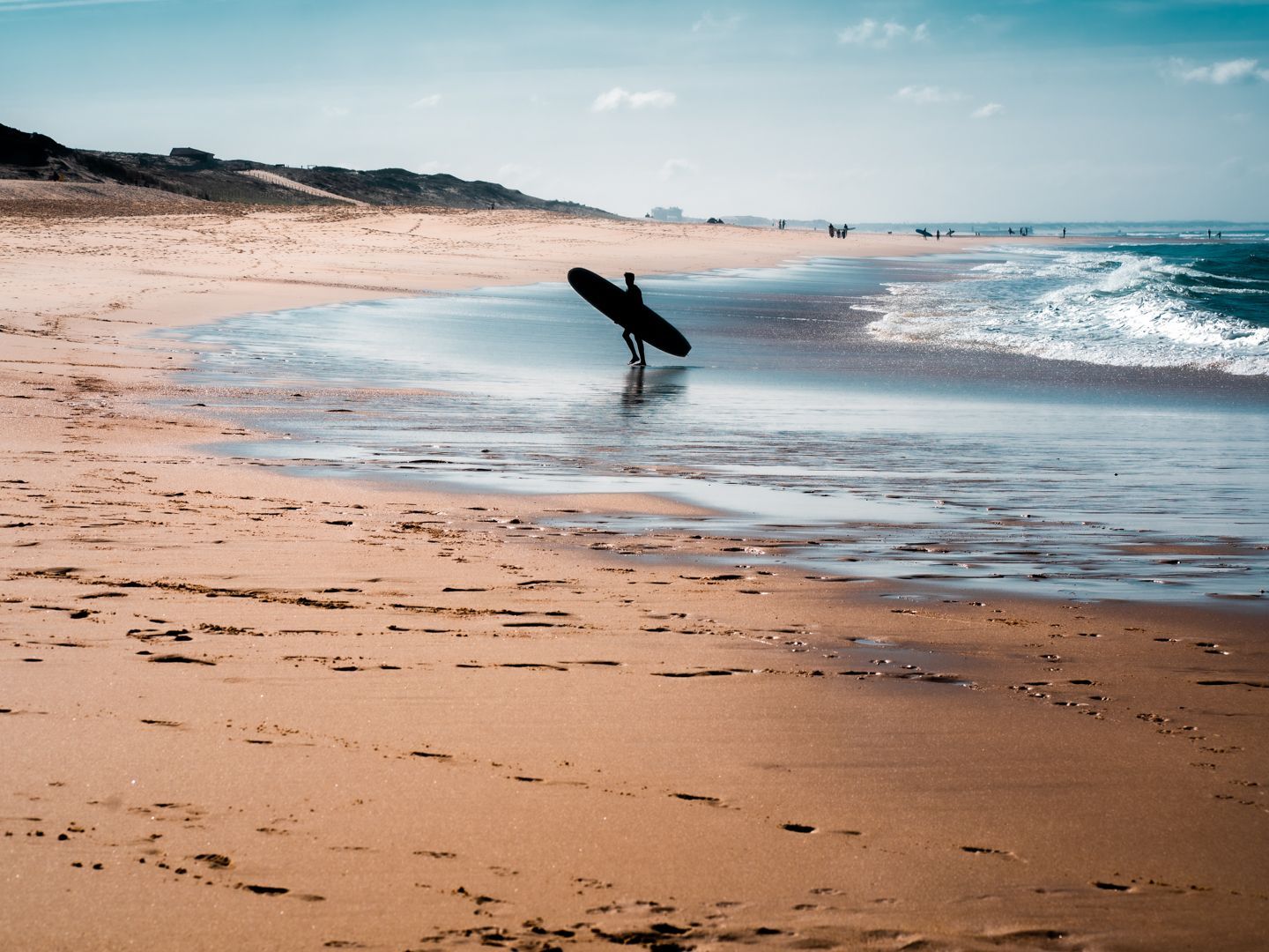 Un surfista in silhouette porta una tavola da surf lungo la riva di una spiaggia sabbiosa mentre le onde si infrangono.