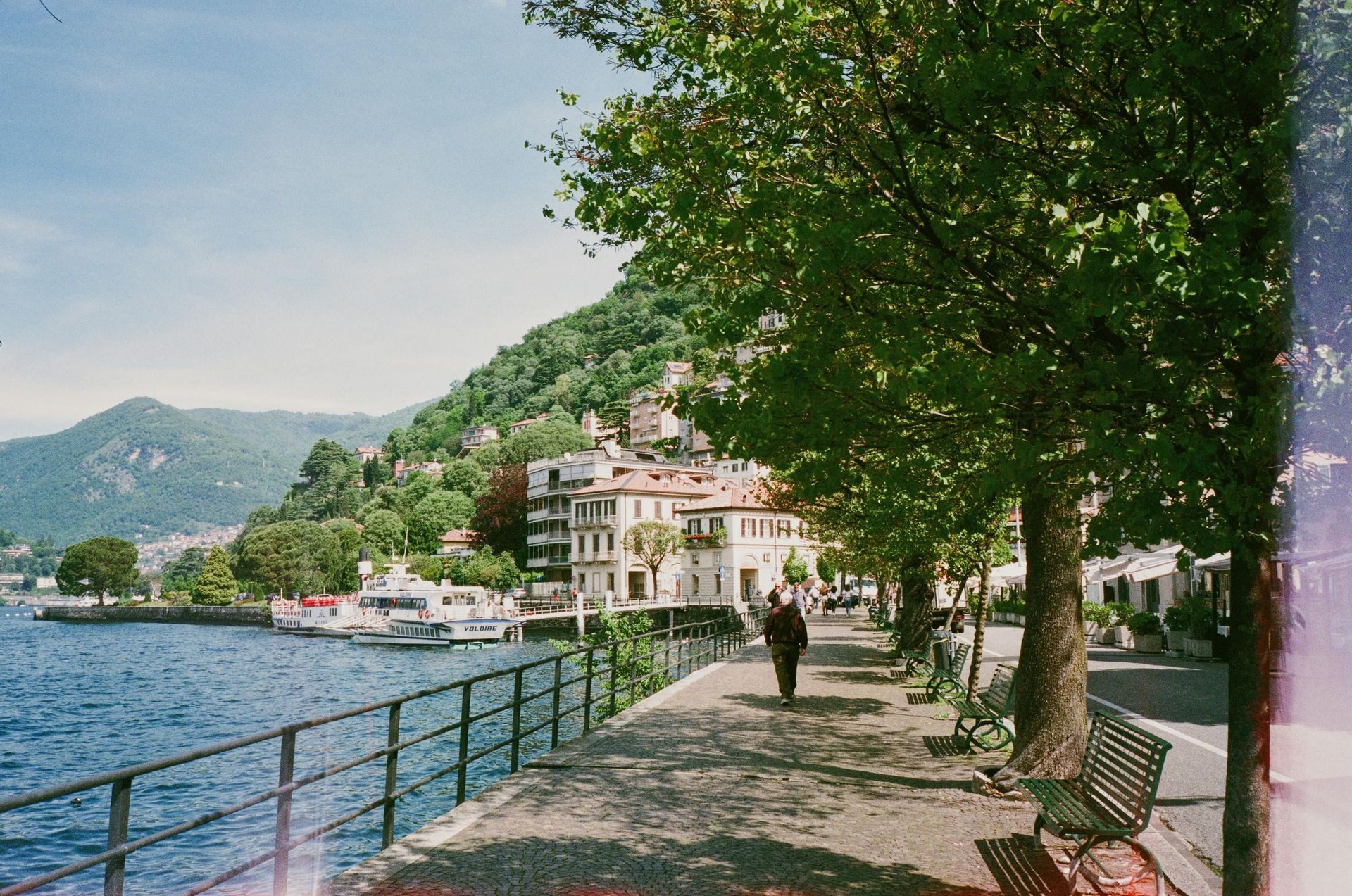 Una persona passeggia lungo un viale alberato e ombreggiato, accanto a un lago con barche ormeggiate e una cittadina collinare.