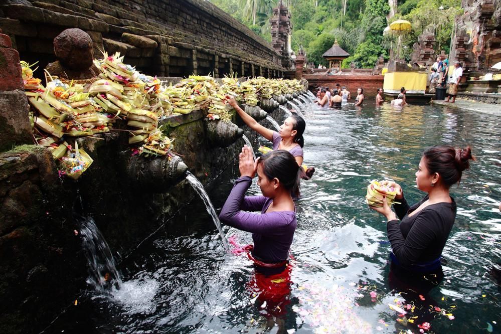 Des personnes effectuent un rituel de purification dans un bassin sacré, priant et déposant des offrandes sous des jets d'eau en pierre.