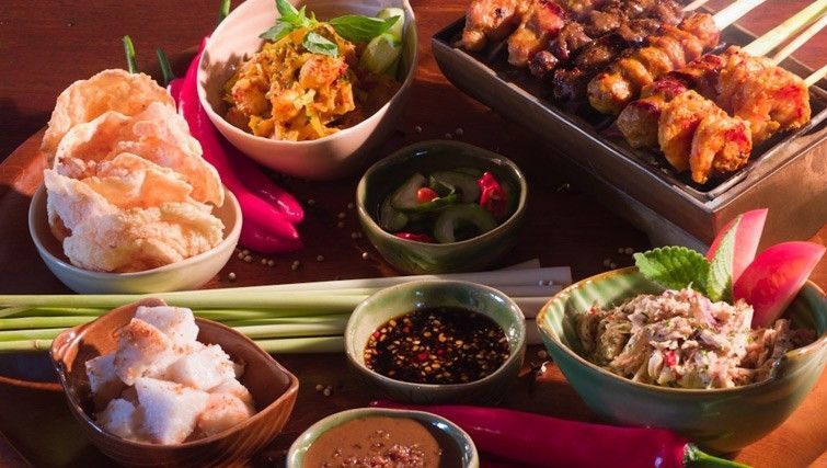Un assortiment de divers plats asiatiques, incluant des brochettes de satay, des crackers, des salades et des sauces pour tremper sur une surface en bois.