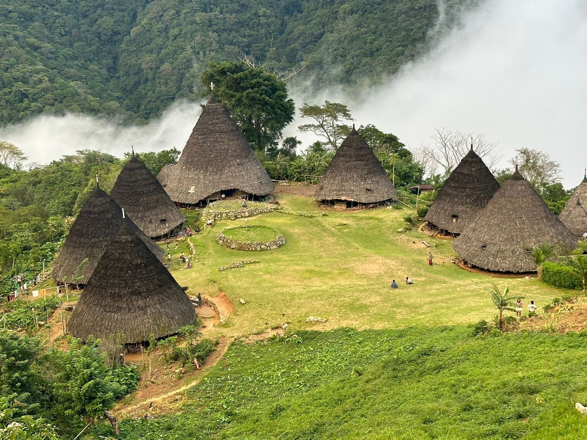 Un village traditionnel avec des huttes coniques aux toits de chaume sur une colline verdoyante, entouré de montagnes boisées et de nuages bas.