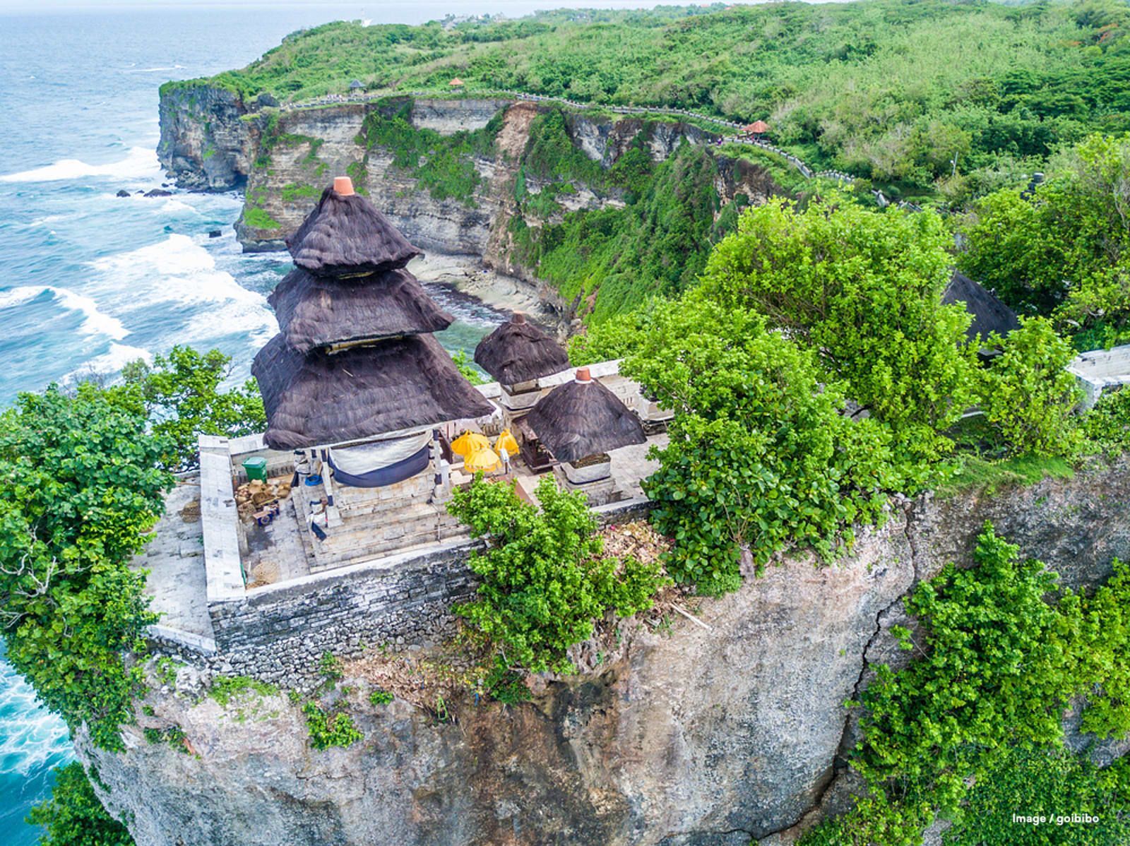 Une vue aérienne d'un temple traditionnel aux toits de chaume, perché sur une falaise verdoyante dominant l'océan.