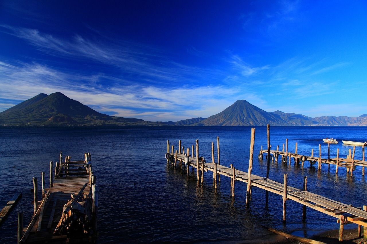 Pontili rustici in legno si estendono su un lago calmo e blu, con grandi montagne visibili attraverso l'acqua sotto un cielo limpido.