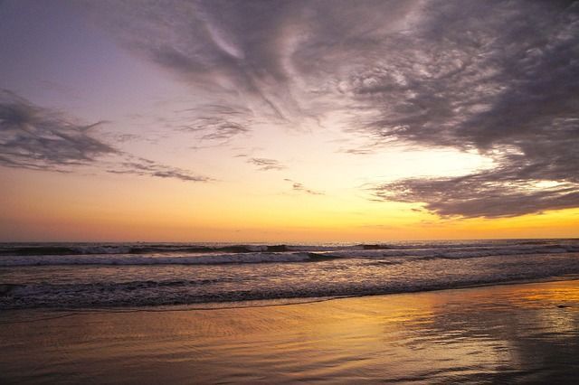 Un tramonto dorato si riflette sulla sabbia bagnata di una spiaggia mentre le onde si infrangono sotto un cielo nuvoloso con sfumature viola.