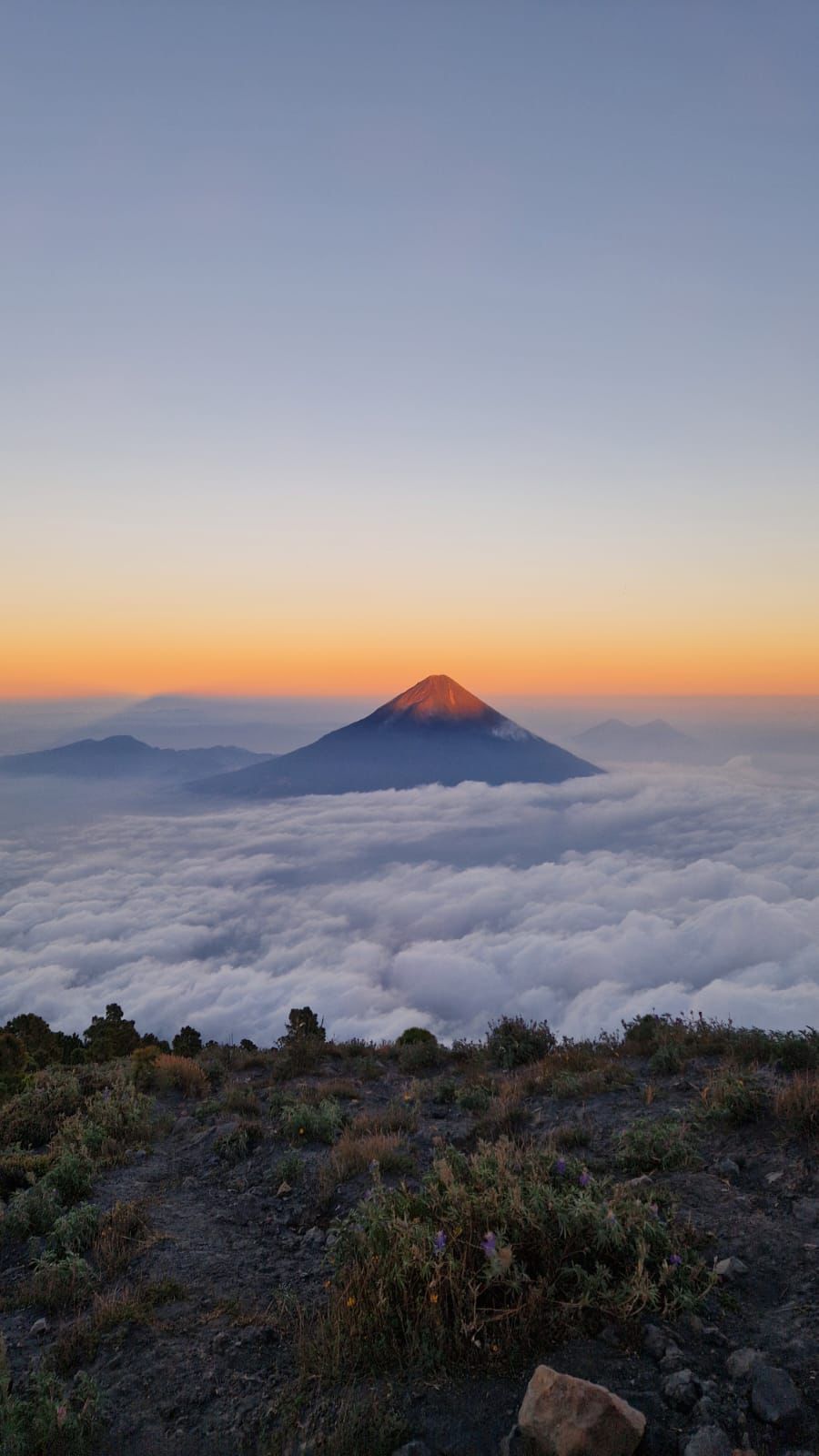 La cima di un vulcano si erge sopra un mare di nuvole, illuminata dalla luce dorata dell'alba, vista da un punto panoramico roccioso.