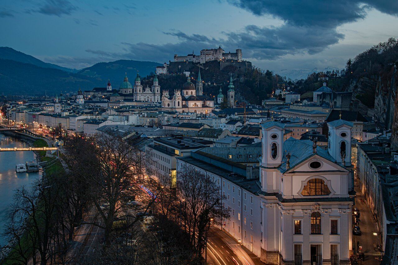 Eine bei Sonnenuntergang beleuchtete Stadtlandschaft mit einer Festung auf einem Hügel, historischen Gebäuden und Bergen in der Ferne unter einem bewölkten Himmel.