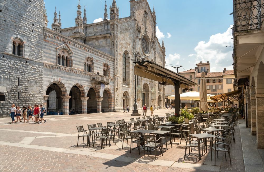 Un caffè all'aperto con tavoli vuoti si trova in una piazza soleggiata e pavimentata di fronte a una grande cattedrale in pietra a strisce.