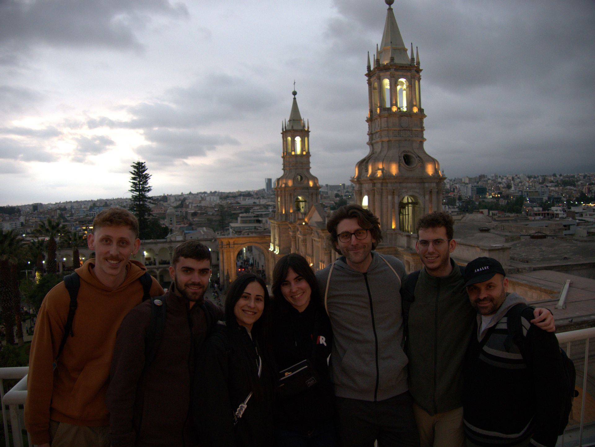 Un viaggio di gruppo WeRoad di sette persone in posa su un punto panoramico con due torri di cattedrale illuminate e uno skyline della città al tramonto.