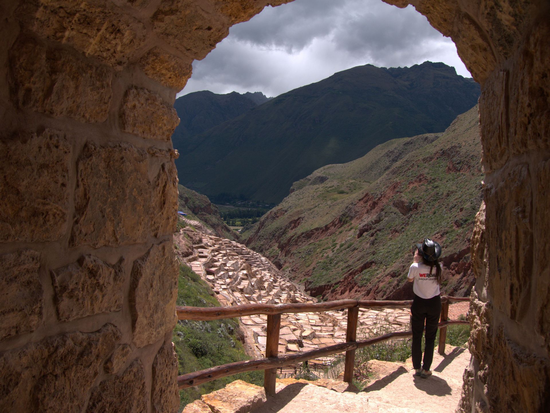 Una persona vista di spalle si trova sotto un arco di pietra, osservando le saline terrazzate in una valle di montagna.