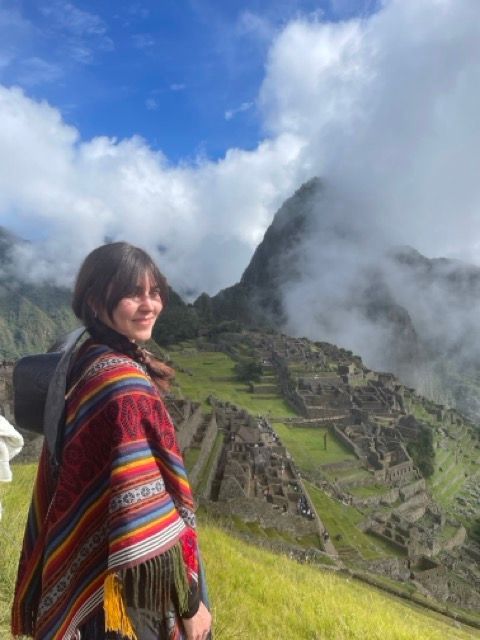 Una donna con un poncho colorato si erge su una collina erbosa che si affaccia su antiche rovine di pietra su un fianco di montagna, sotto un cielo nuvoloso.