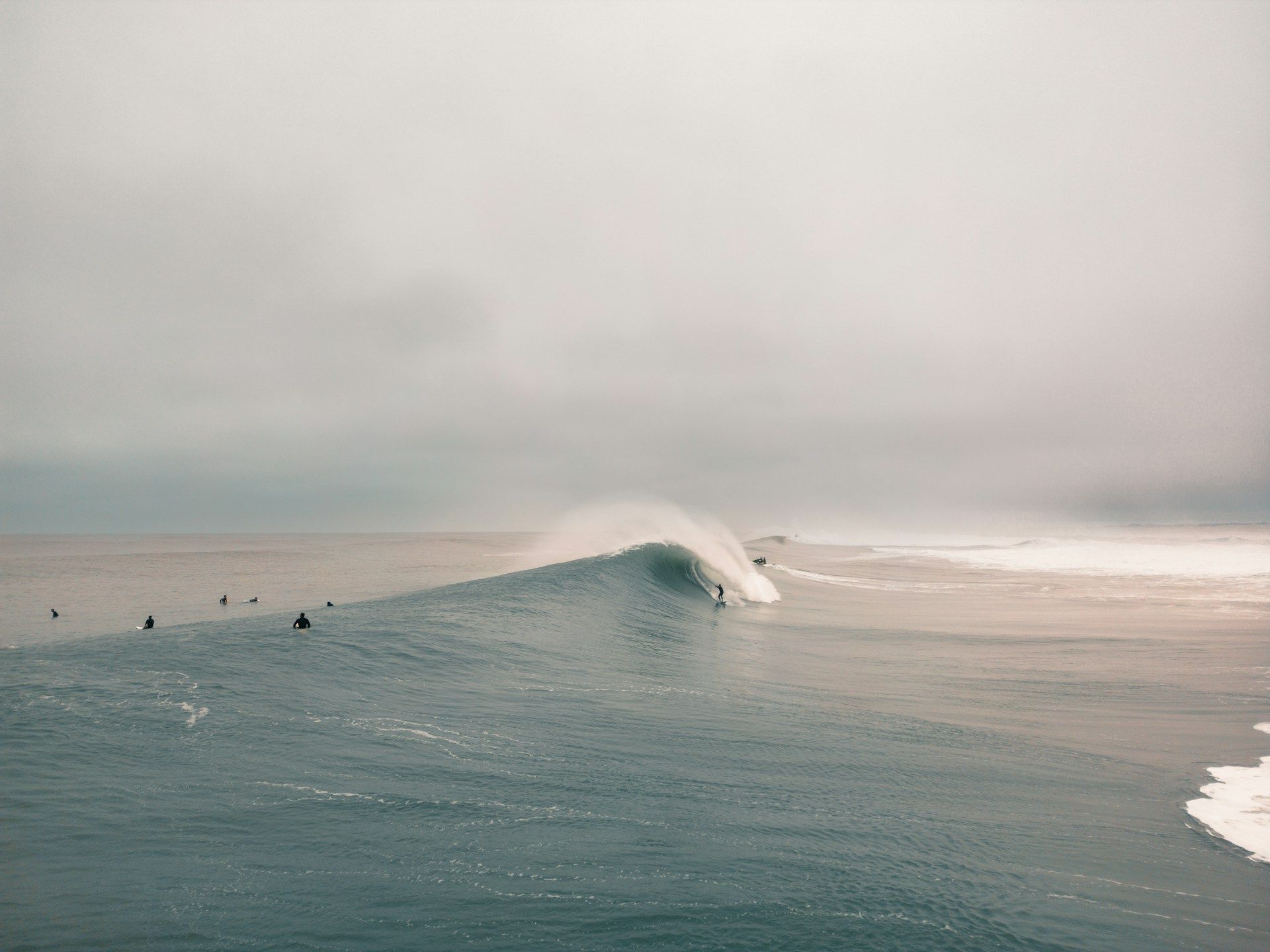 Un surfista cavalca una grande onda tubante nell'oceano, mentre altri surfisti attendono sotto un cielo nuvoloso.
