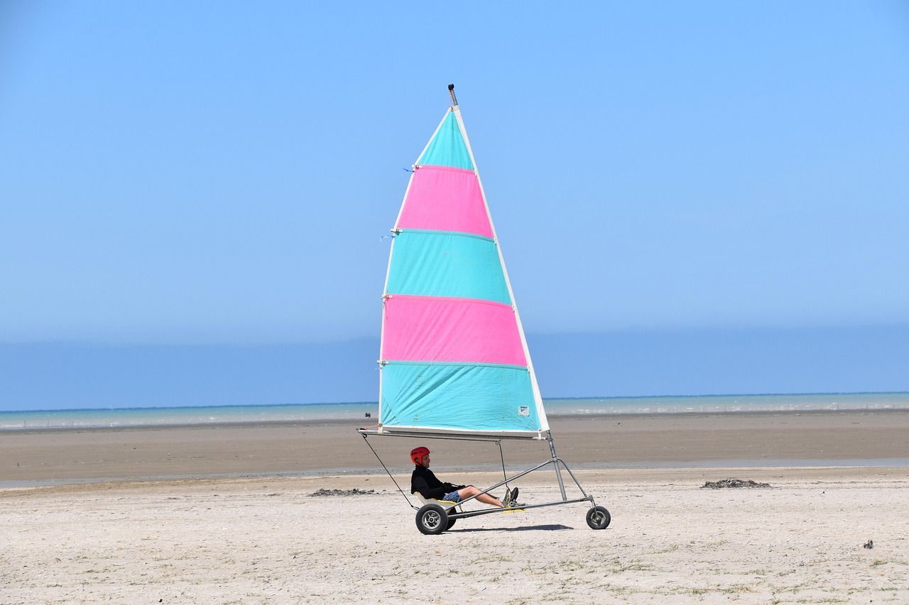 Une personne dans un char à voile avec une voile rose et turquoise est assise sur une large plage de sable près de l'océan sous un ciel bleu clair.